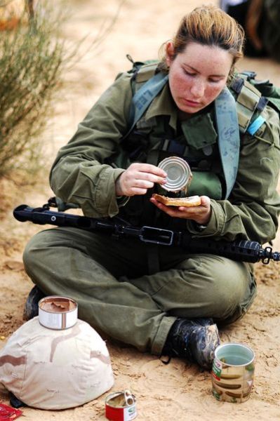 以色列女兵在享用野战口粮，貌似军靴旁边的大罐头是蔬菜罐头，头盔上和头盔旁的罐头是肉罐头。正在往面包上倒的是金枪鱼罐头。